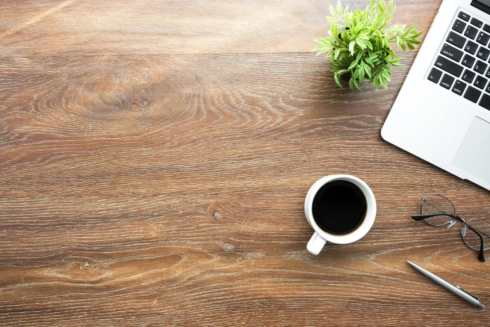 Plan de travail en bois vu de dessus, coin d’ordinateur portable, tasse de café noir, lunettes et petite plante verte