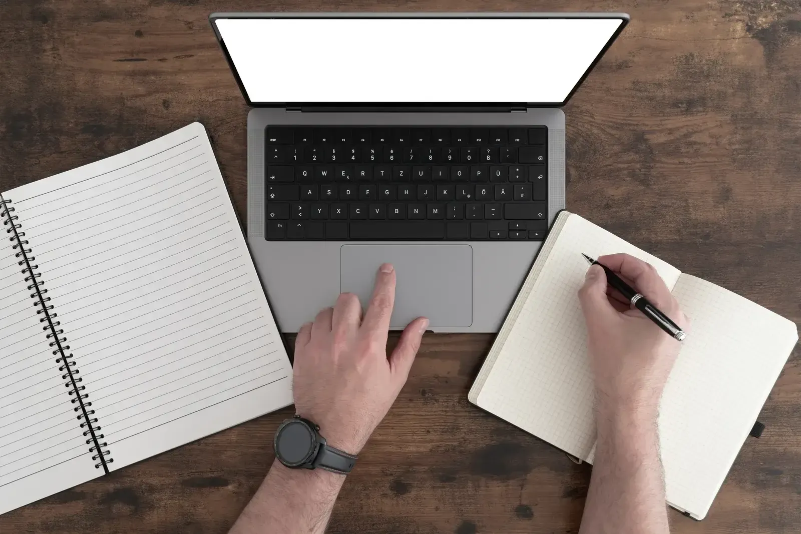 Vue de dessus d’une table en bois foncé : une main sur le clavier d’un ordinateur portable, l’autre écrivant dans un carnet ouvert, un bloc à spirale posé à côté