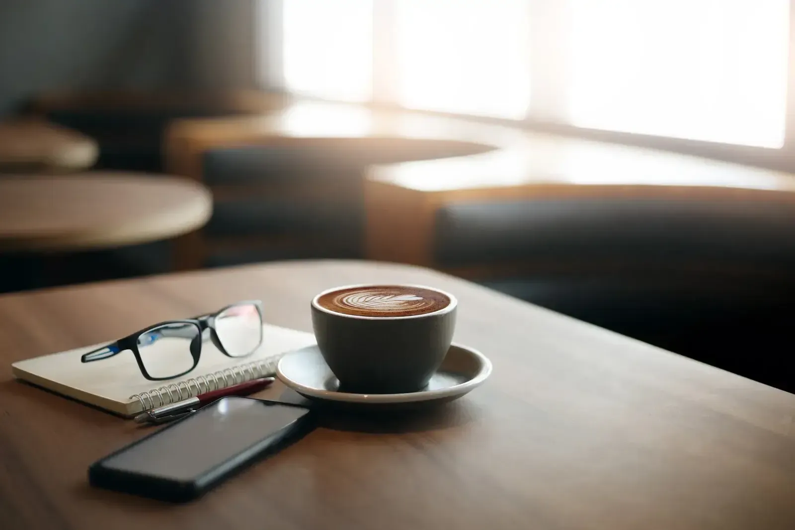 Carnet à spirale, lunettes, stylo, téléphone et tasse de café posés sur une table en bois devant une fenêtre lumineuse
