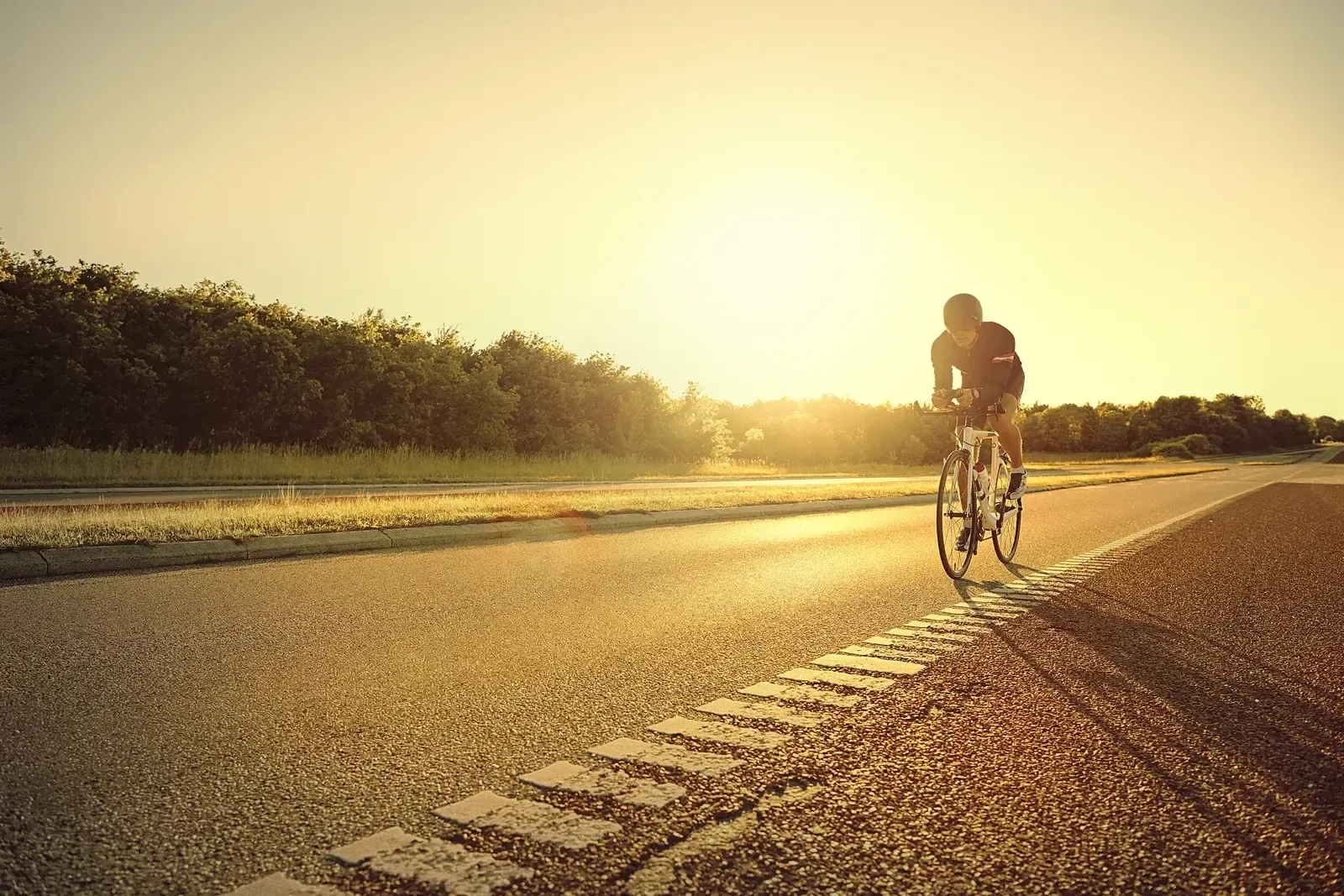 Un cycliste seul remonte une route déserte face au soleil levant, la chaussée et les talus baignés d’une lumière dorée