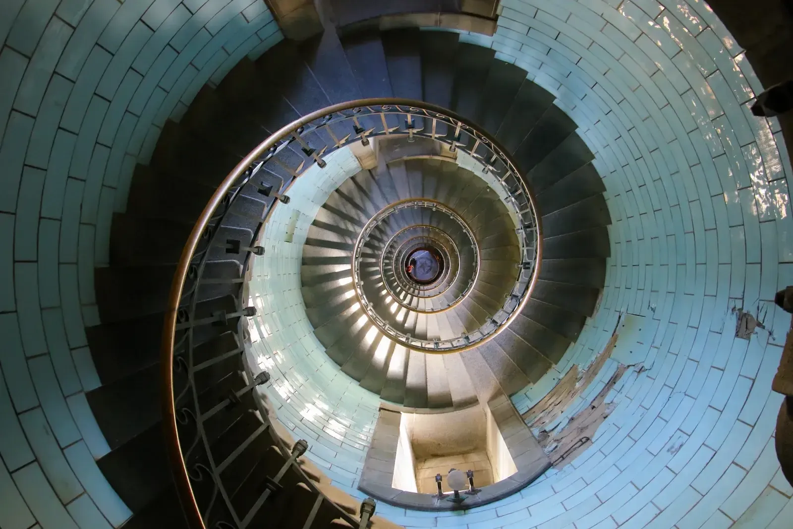 Vue plongeante sur l’escalier en colimaçon d’un phare, les marches s’enroulant jusqu’à un point minuscule entre des murs revêtus de faïence turquoise