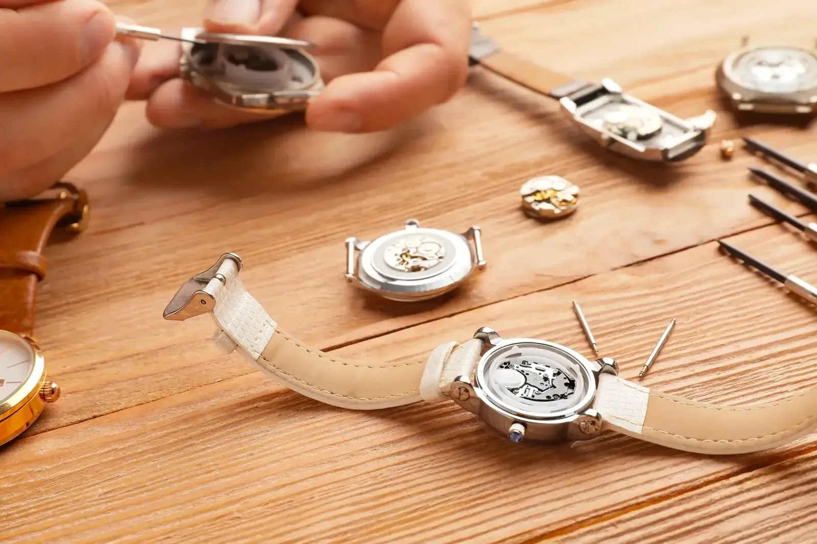 Les mains d’un horloger ouvrent le boîtier d’une montre, plusieurs montres démontées et de fines pincettes posées sur un établi en bois clair