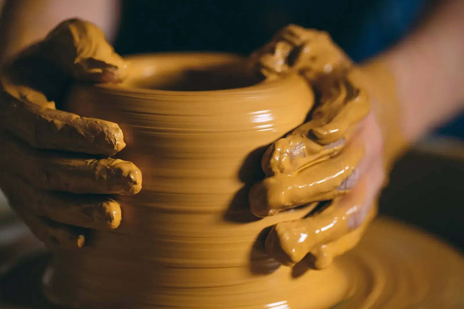 Mains couvertes d’argile qui façonnent un pot sur un tour de potier, lumière ambrée d’atelier