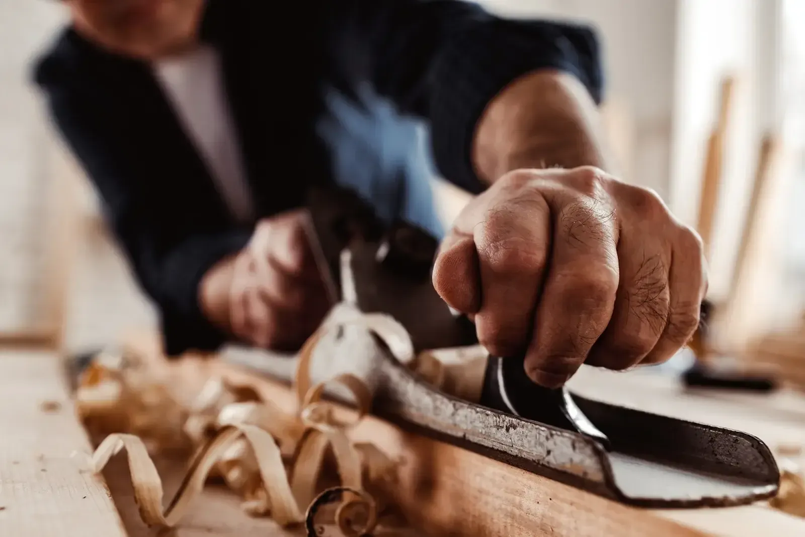 Mains d’un menuisier qui poussent un rabot sur une planche de bois, copeaux enroulés sur l’établi
