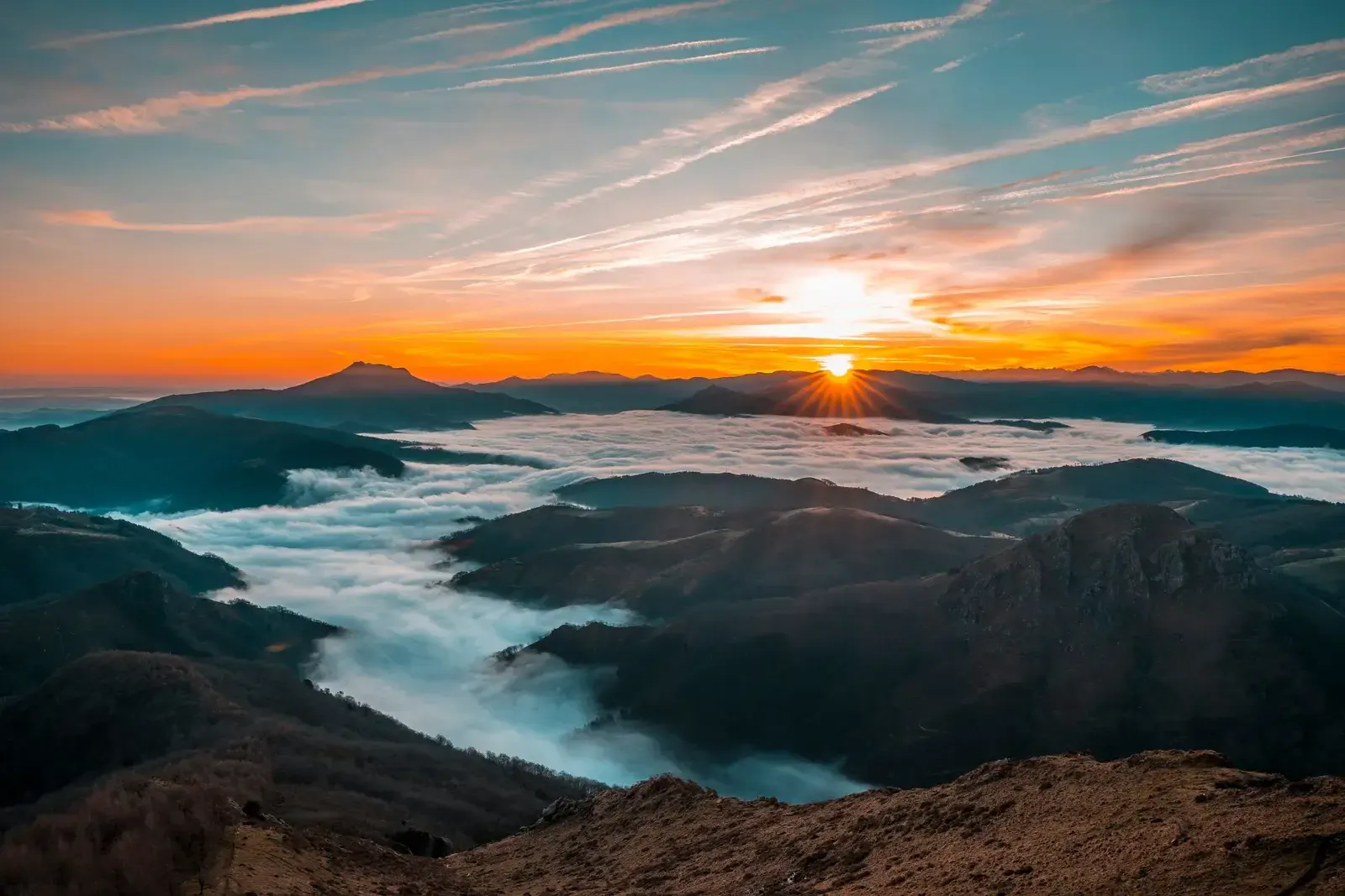 Sommets qui émergent d’une mer de nuages au lever du soleil, ciel orangé au-dessus des crêtes