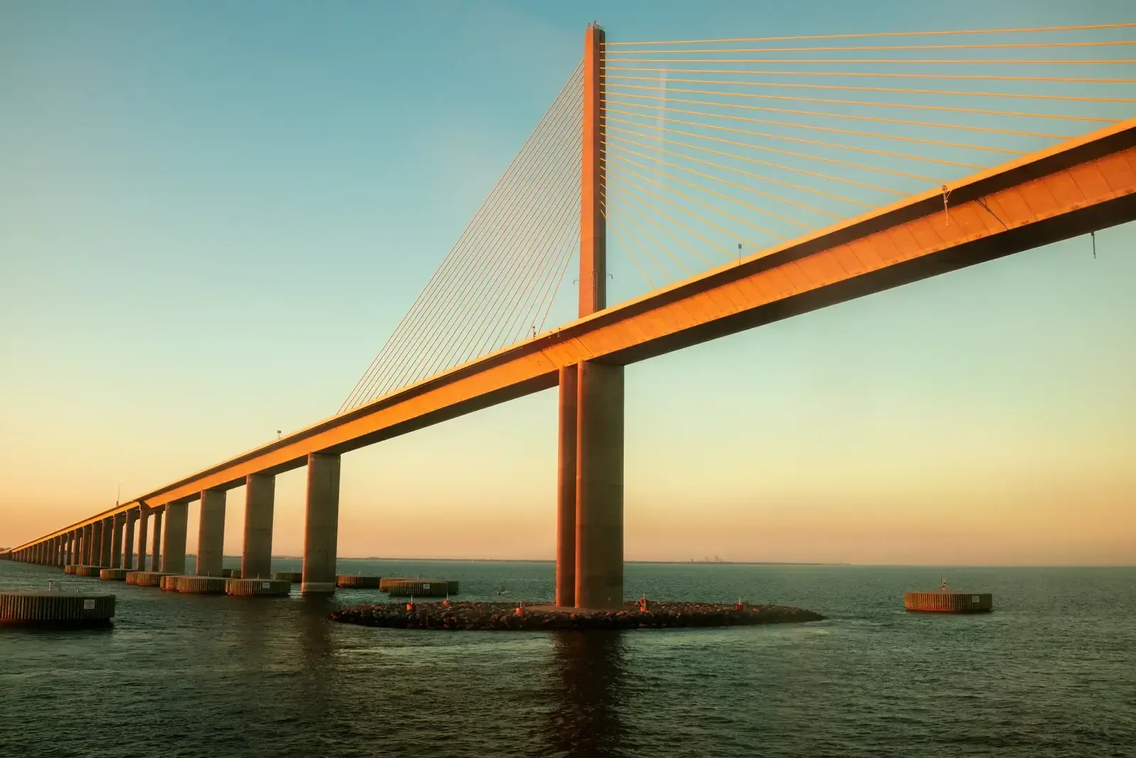 Long pont à haubans au tablier orangé s’étirant vers l’horizon au-dessus d’une mer calme, sous un ciel sarcelle et doré