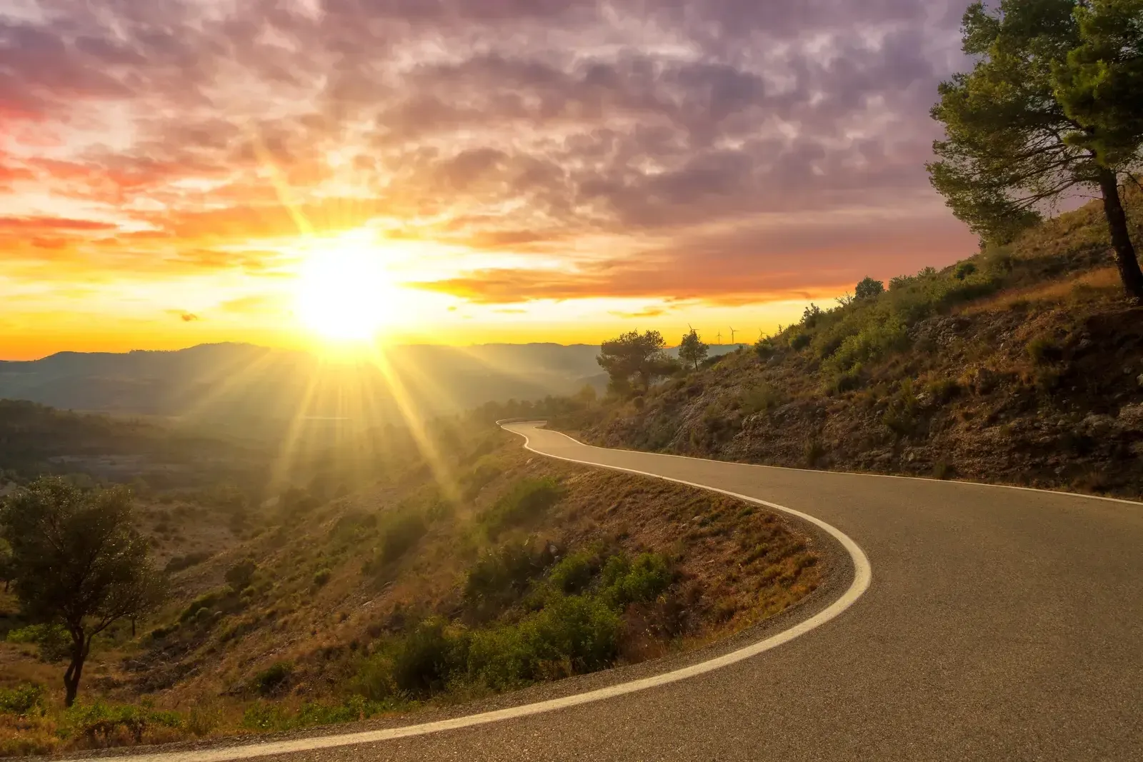 Route sinueuse traversant des collines sèches, le soleil bas à l’horizon éclairant un ciel orangé et nuageux