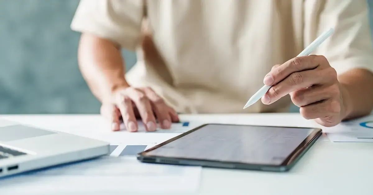 Main tenant un stylet au-dessus d'une tablette posée près d'un ordinateur portable, sur un bureau clair