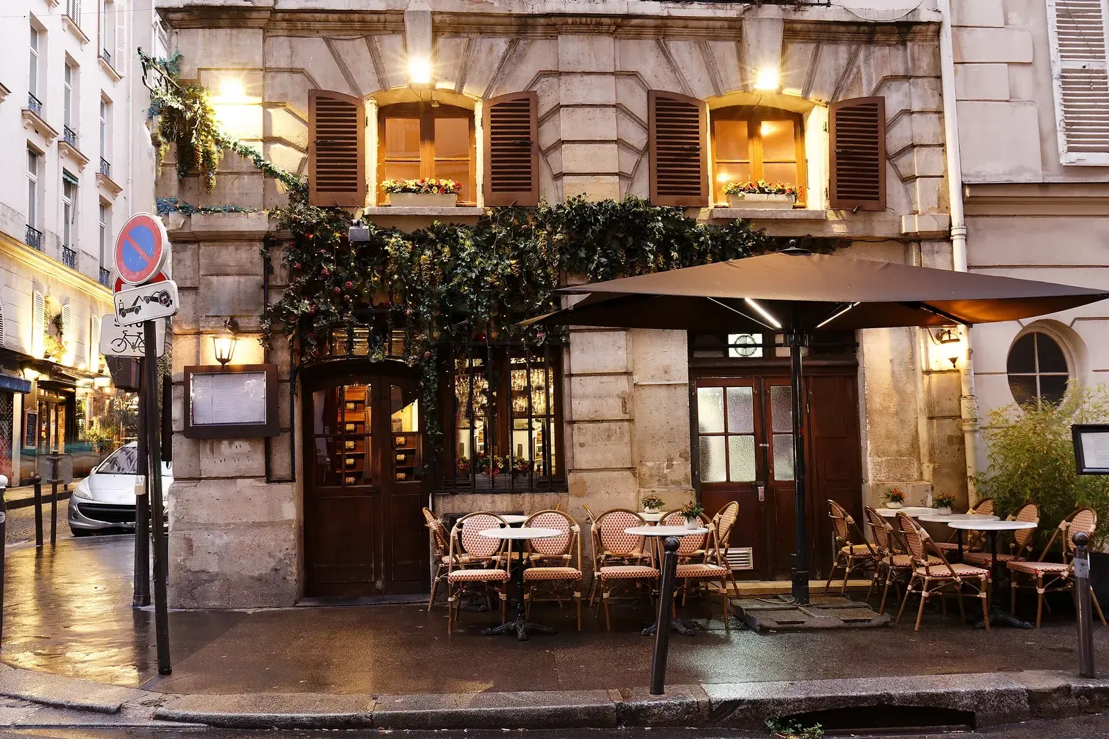 Devanture d’un restaurant à la tombée du jour, fenêtres à volets éclairées de jaune et tables de terrasse sous un parasol