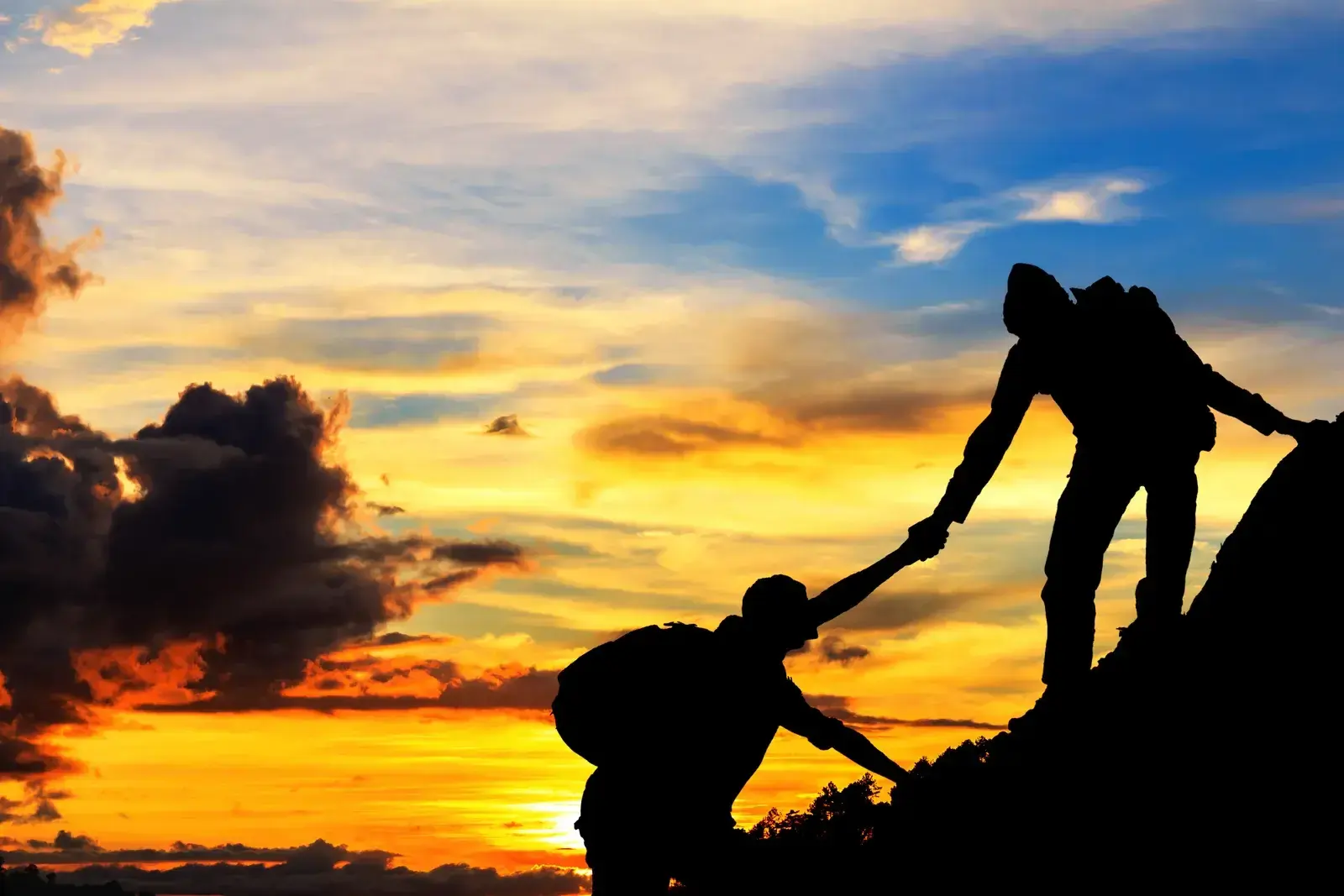 Two hikers in silhouette on a rocky ridge, one reaching a hand down to help the other up, against an orange sunset sky