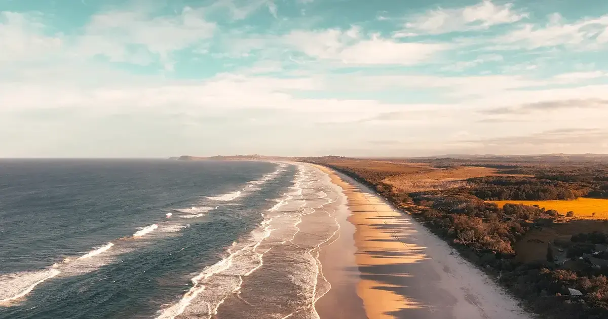 A long beach seen from the air, parallel trains of swell running in onto the sand in the morning haze