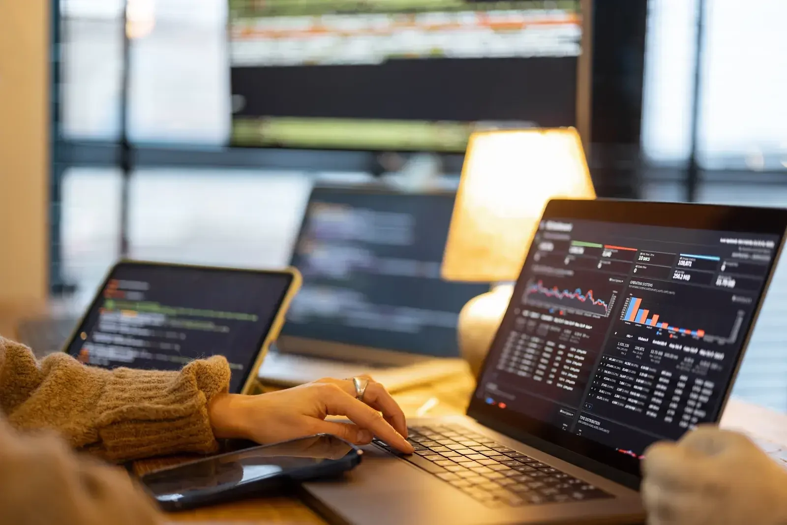 Hands of a person in a beige sweater on a laptop keyboard, several screens showing graphs and dashboards, a warm lamp in the background