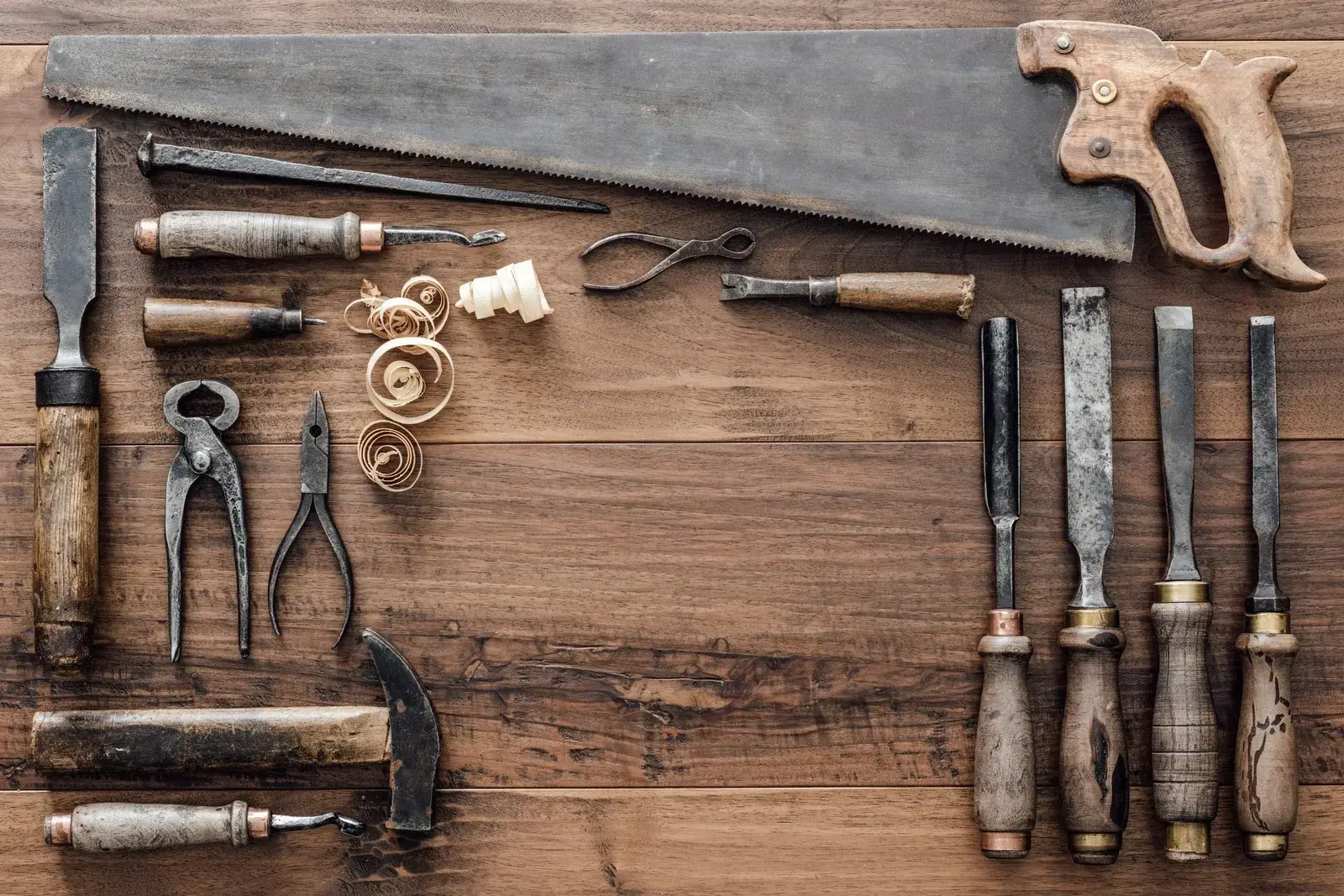 An old saw, wood chisels, pincers and a hammer laid side by side on a pale wooden workbench