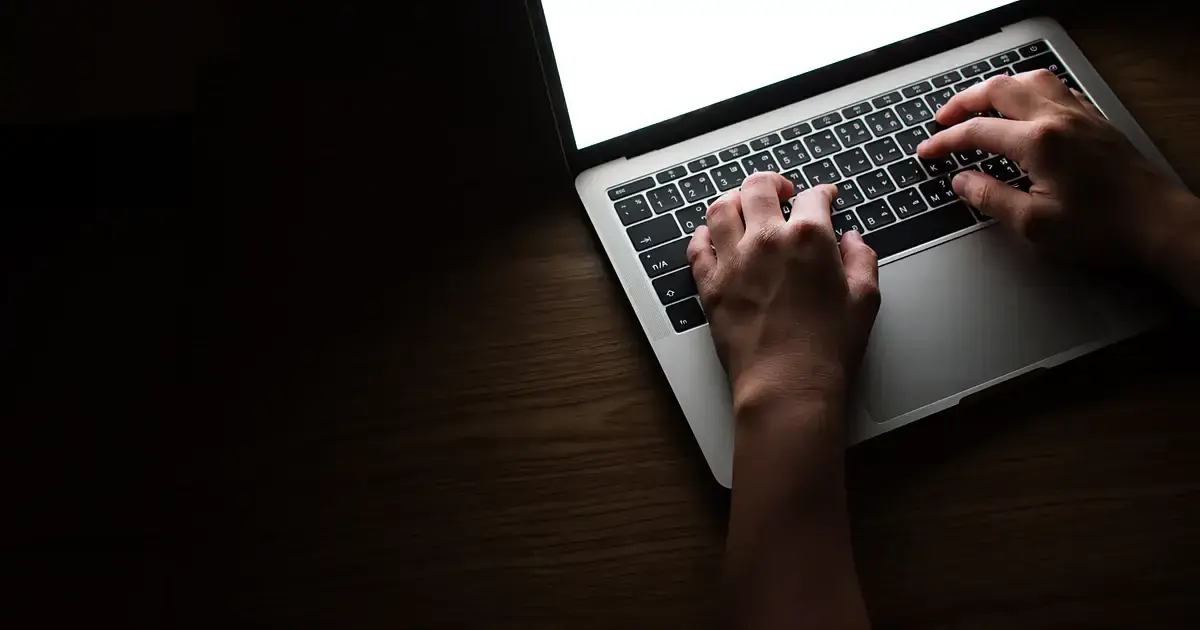 Mains posées sur le clavier d'un ordinateur portable dans une pièce sombre, lumière chaude rasante sur le bois