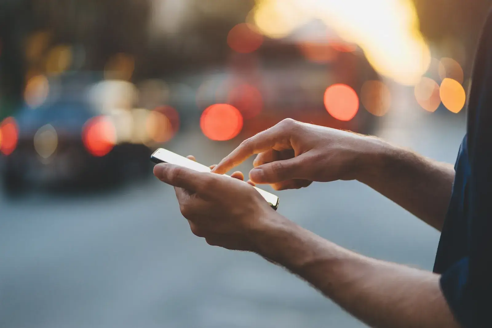Deux mains tiennent un téléphone dans une rue le soir, halos orange des phares de voitures en arrière-plan