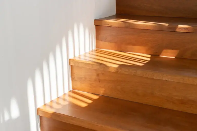 Escalier en bois clair dont les marches successives sont striées de bandes de lumière dorée le long d’un mur blanc