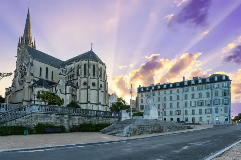 Église en pierre claire et immeubles du centre historique de Pau au lever du soleil, rayons dorés dans un ciel violet